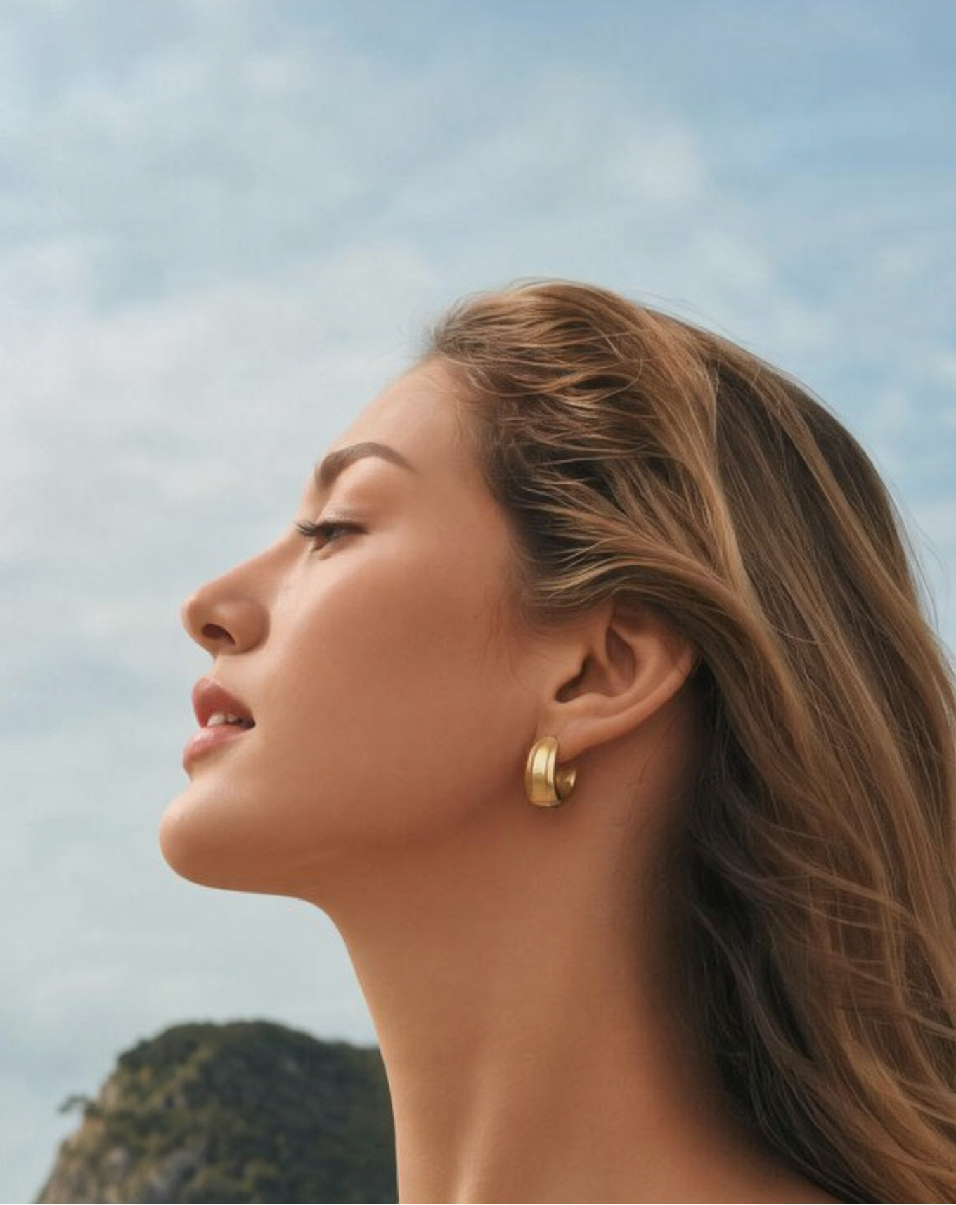 Moska Cosmetics model wearing gold hoop earrings with a blurred natural background with lip plumper and radiance pocket blush